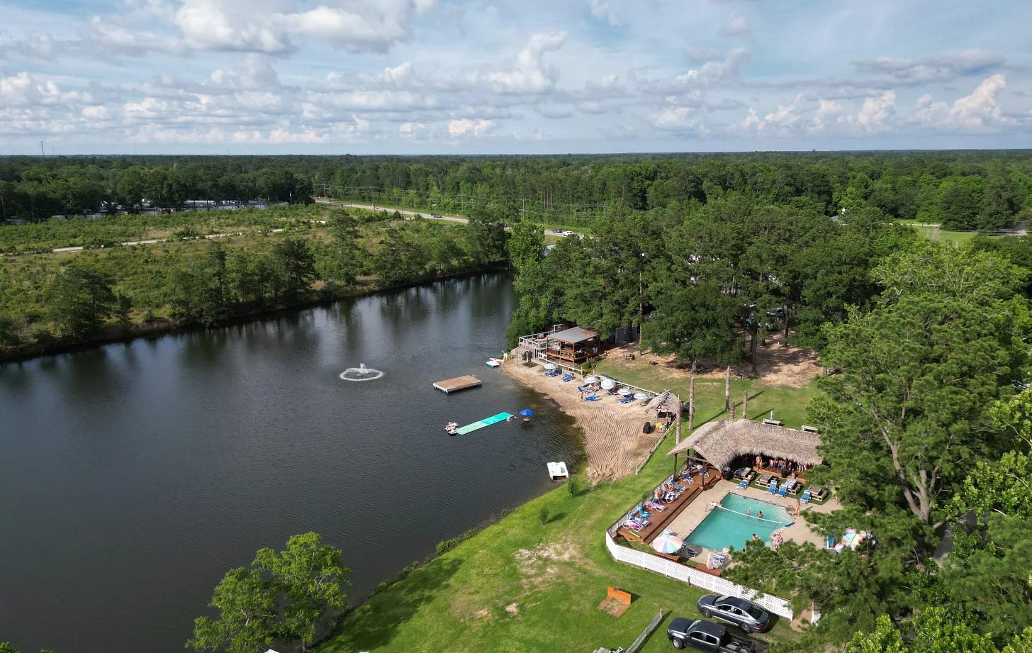 An aerial view of Emerald Lake Resort. A dark, tree-lined lake fills the left of the frame, with a fountain spraying near a floating dock and a green water mat. Along the right shore sits a sandy beach with lounge chairs, a thatched-roof pool area where people swim and sit poolside, a two-story deck building among pines, and parked cars on a grassy lot. Beyond the resort, a road runs past a large cleared field before dense forest stretches to the horizon under a blue sky with scattered clouds.