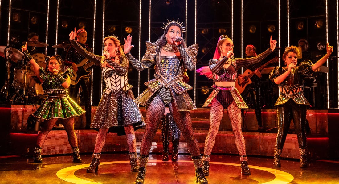 Five women stood on stage wearing sparkling dresses and fish nets, singing into microphones