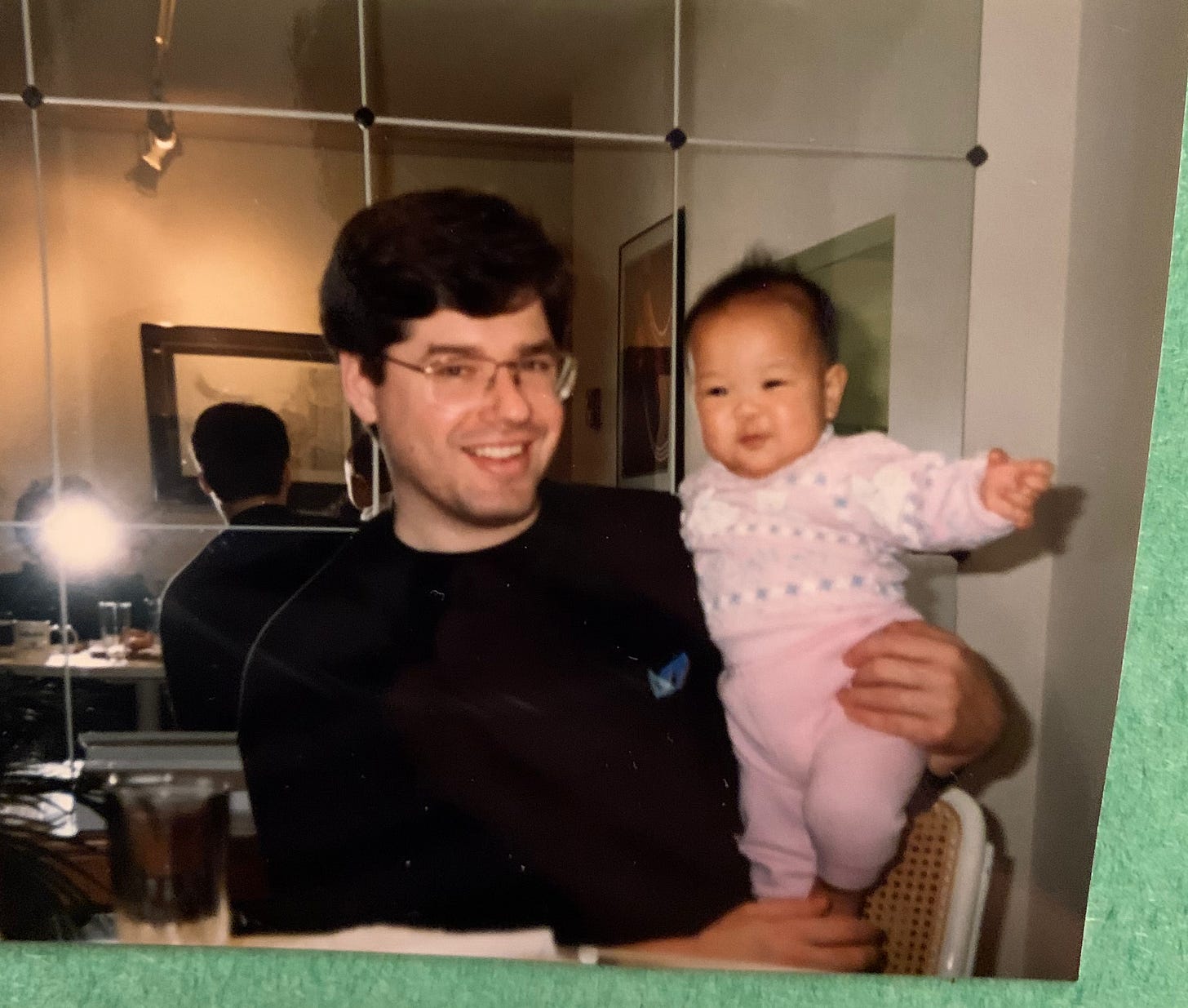 Uncle Richard sits at a dining table, holding baby me again. At his back is a wall of mirrors. He's wearing a black crewneck sweater and the same rectangular glasses, and I'm wearing a little matching pink knitted set with a white and light green pattern on the sweater. We're both smiling. Circa December 1991.