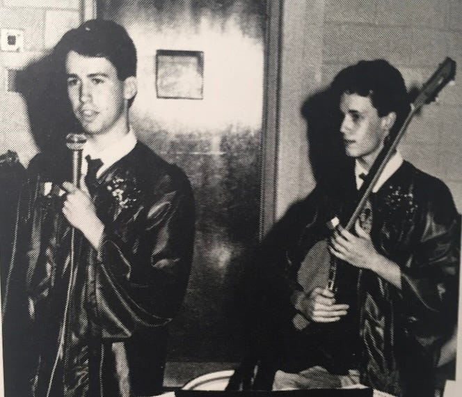 Two high school kids in graduation smocks, one with a microphone and one with a banjo