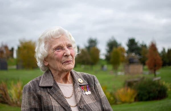 A woman with white hair wears a blazer festooned with medals, in a rural setting. 