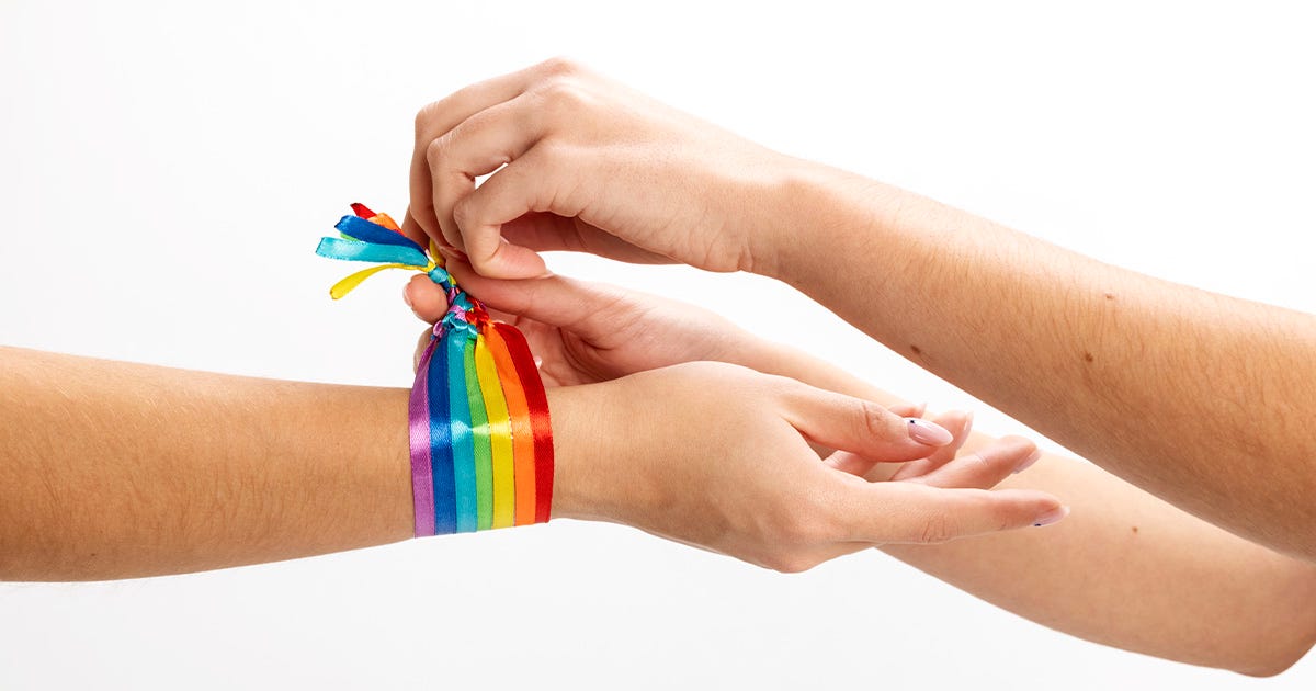 Imagem de dois braços que se encontram, sendo que um deles amarra no pulso do outro uma pulseira multicolorida, que faz alusão ao universo LGBTQIA+ Imagem de dois braços que se encontram, sendo que um deles amarra no pulso do outro uma pulseira multicolorida, que faz alusão ao universo LGBTQIA+