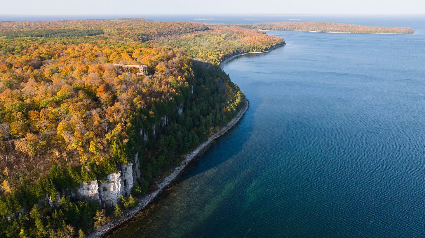 A wooden boardwalk along the cliffs of Door County, Wisconsin, surrounded by glowing amber and red autumn trees reflected in Lake Michigan.