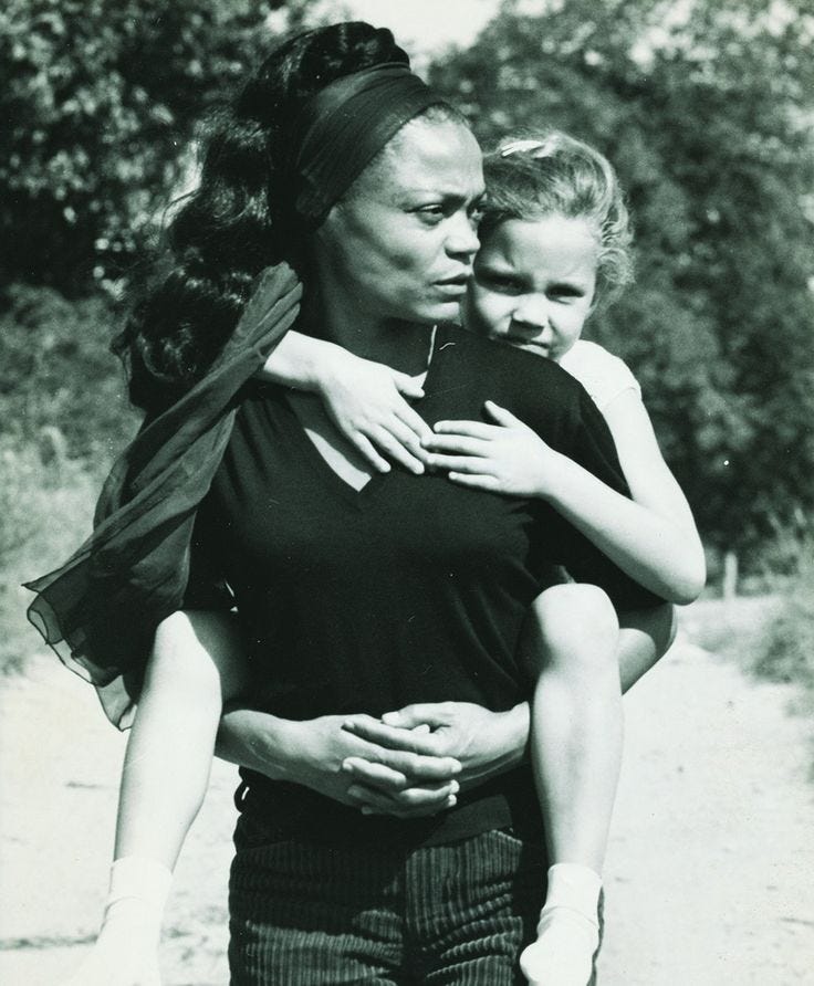 This may contain: a woman holding a child in her arms on a dirt road with trees in the background
