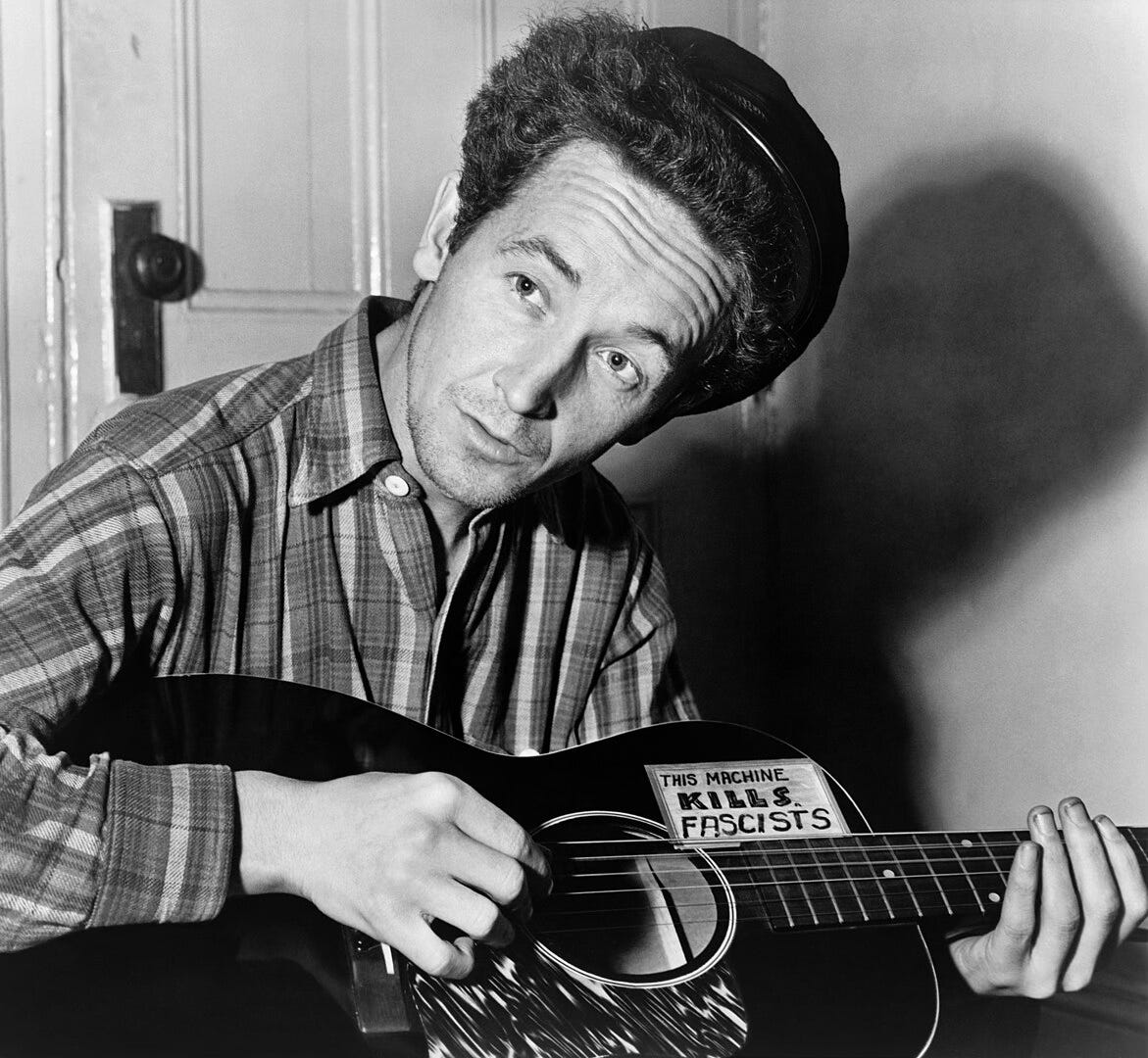 Woody Guthrie holding a guitar emblazoned with a sticker that says "This machine kills fascists."