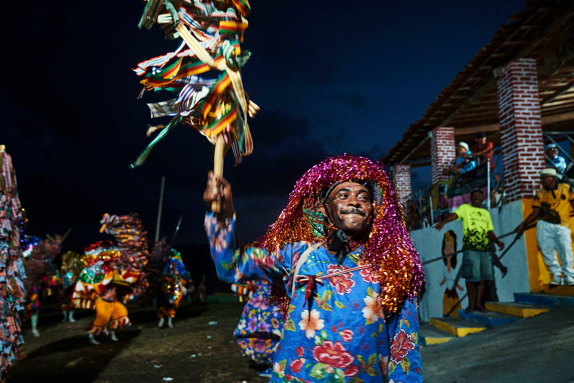 A smiling man in a colorful costume and shiny tinsel wig waves a decorated stick during a lively outdoor festival at night, with other dancers and spectators visible in the background.