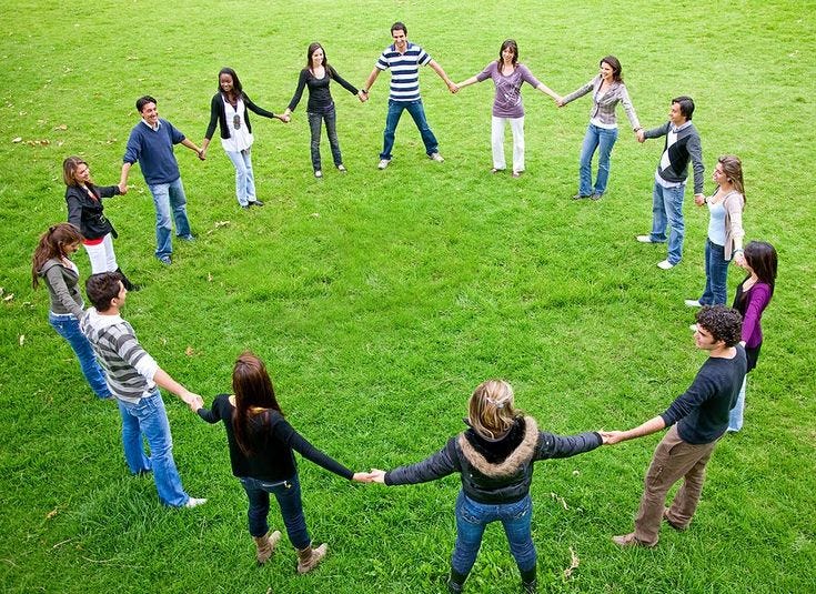 This may contain: a group of people holding hands in the middle of a circle on grass with trees in the background
