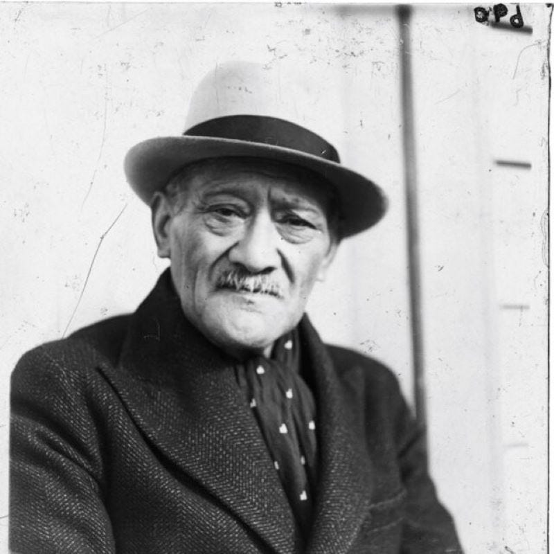 A black-and-white portrait of Tahupotiki Wiremu Ratana taken by Sam Dale. It shows T. W. Ratana, an old Māori man with a moustache, from the chest up, wearing a black coat, a black scarf with white dots and a Panama or Homburg hat.