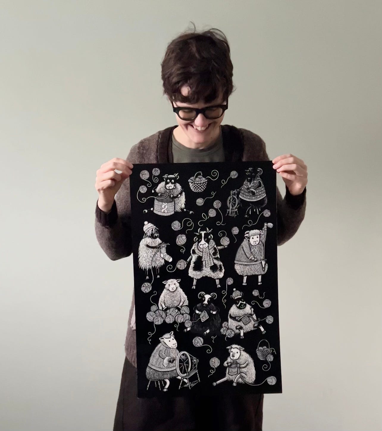 Katie stands against a green wall holding a large piece of scratchboard art, showing sheep knitting, crocheting and spinning