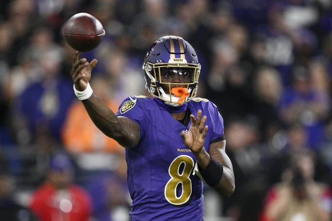 Lamar Jackson wearing a purple Baltimore Ravens jersey with number 8 stands on a football field holding a football in his right hand while raising his left arm in a throwing motion his face is partially visible through the facemask of his helmet with orange accents the background shows a blurred stadium crowd and field during a game.