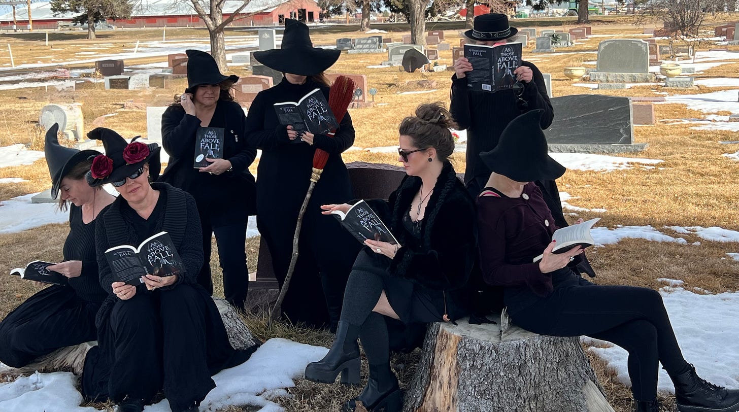 Seven women dressed as witches reading a book called As Those Above Fall in a cemetery
