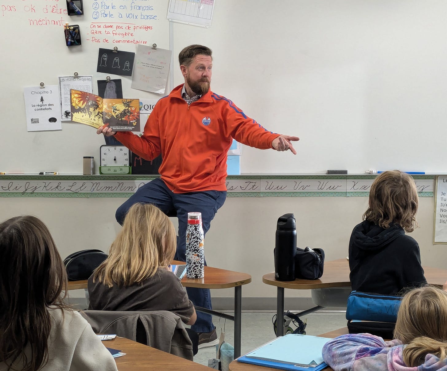MLA Kyle Kasawski is sitting in front of a group of students in a classroom wearing an orange Oilers sweater and reading a book.