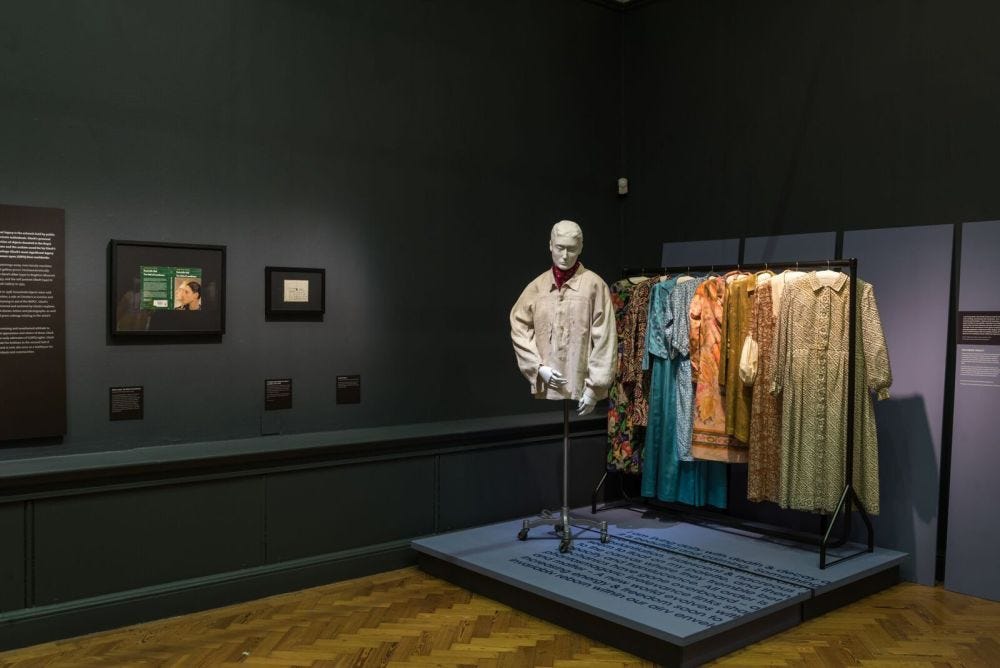 Museum exhibit showcases a white mannequin in a white smock beside colorful vintage dresses on a black rack. Dim lighting and framed art add a reflective tone. There is a plaque with text on the far left of the wall, and text plaques underneath each work. There is also black text on the blue platform that Gluck's clothes are on. The walls are black and floors are light and medium brown wood. Museum exhibit showcases a white mannequin in a white smock beside colorful vintage dresses on a black rack. Dim lighting and framed art add a reflective tone. There is a plaque with text on the far left of the wall, and text plaques underneath each work. There is also black text on the blue platform that Gluck's clothes are on. The walls are black and floors are light and medium brown wood.