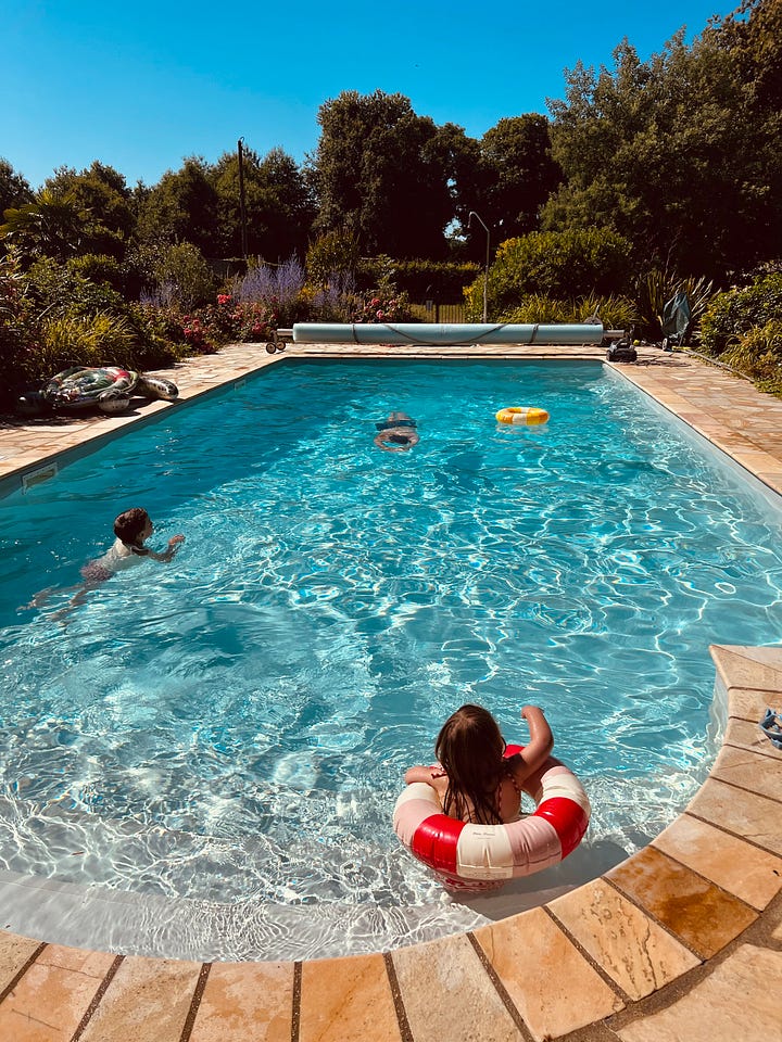 Two photos side by side. On the left, three children swim in a rectangular in-ground pool ringed by stone paving and flowering shrubs, one floating in a red-and-white ring float. On the right, two children nap on a beige sofa against an exposed stone wall hung with framed portraits, a red blanket draped behind them.