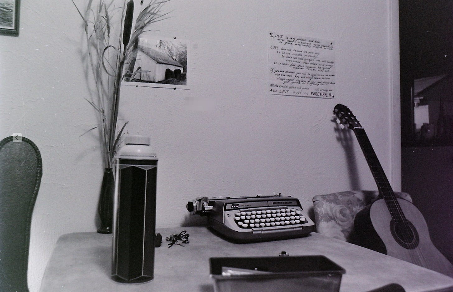 A black-and-white photograph of a quiet room corner with a vintage typewriter on a table, a classical guitar leaning against a chair, and a tall thermos in the foreground. On the wall above the typewriter are a photo of a barn and a handwritten note or poem about love. A tall vase with dried reeds stands next to the wall, and a pair of keys rests on the table. The scene evokes a nostalgic, creative atmosphere.