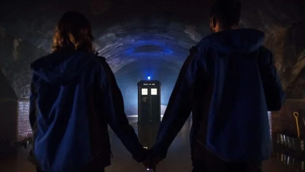 This image shows Lin (played by Charlotte Ritchie, left, back to camera) holding hands with Mitch (played by Nikesh Patel, right, back to camera) as the TARDIS's lamp flashes blue in the distance. The TARDIS is perfectly centered between their bodies. This image shows Lin (played by Charlotte Ritchie, left, back to camera) holding hands with Mitch (played by Nikesh Patel, right, back to camera) as the TARDIS's lamp flashes blue in the distance. The TARDIS is perfectly centered between their bodies.