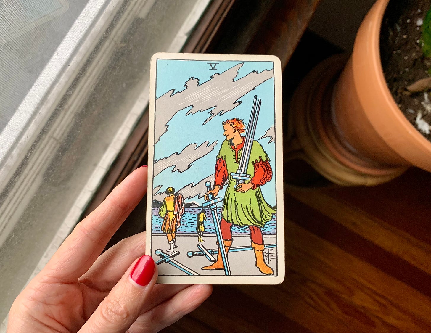 A hand is holding a tarot card, five of swords by pamela colman smith for the Rider-Waite-Smith tarot. In the image, a person in the foreground is holding three swords and two are on the ground. There are two people with their backs turned to them in the background. They are looking back toward the others. Jagged clouds fill the sky. Behind the card is a window sill, a plant in a terra cotta pot and a wood floor.
