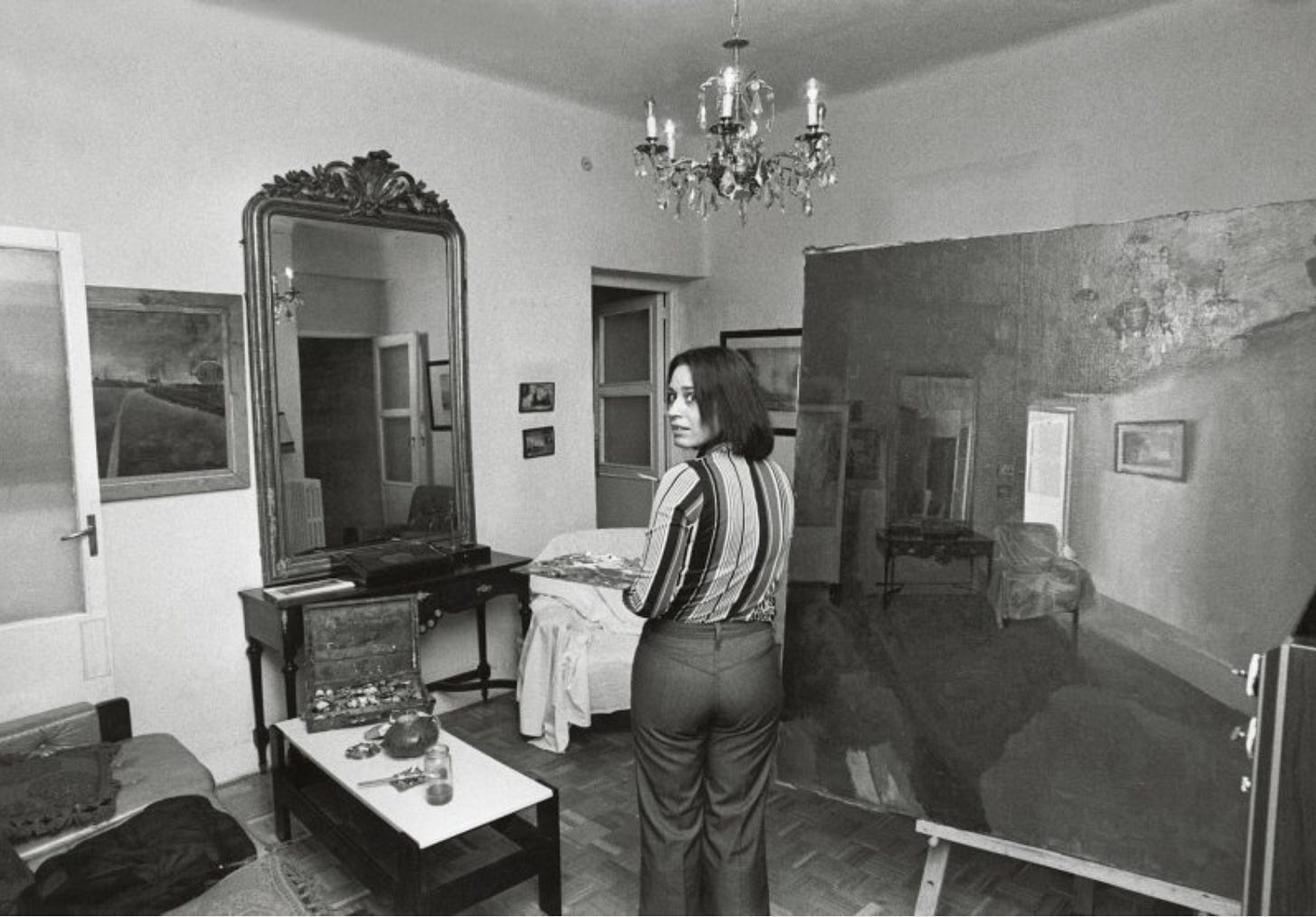 Fotografia in bianco e nero che mostra Isabel Quintanilla in piedi nel suo studio, rivolta verso la macchina fotografica. È davanti a una grande tela che rappresenta un interno domestico. Intorno a lei ci sono mobili antichi, un grande specchio e oggetti da pittura su un tavolino. La stanza è luminosa, con porte e quadri alle pareti.