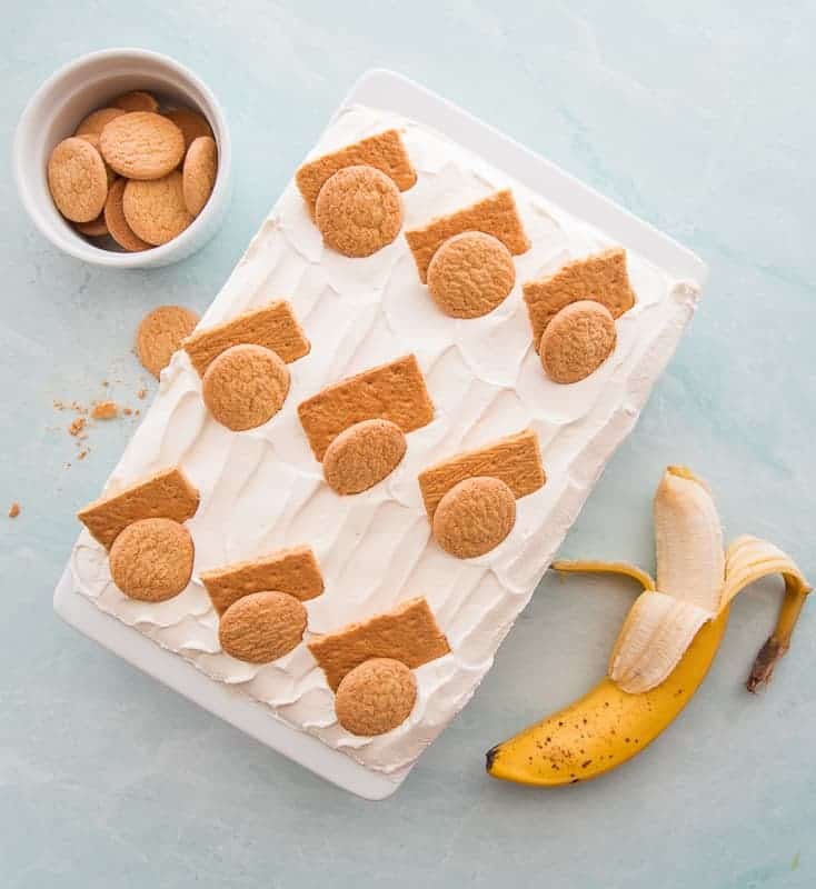An overhead image of the Homemade Banana Pudding. In a white ceramic baking dish and garnished with graham cracker pieces and vanilla wafers. A partially peeled banana sits at bottom right. A white bowl filled with vanilla wafers is in the top left corner. An overhead image of the Homemade Banana Pudding. In a white ceramic baking dish and garnished with graham cracker pieces and vanilla wafers. A partially peeled banana sits at bottom right. A white bowl filled with vanilla wafers is in the top left corner.