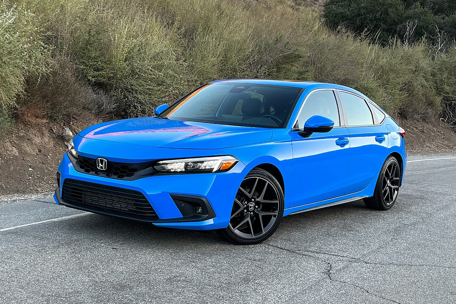 A Boost Blue 2024 Honda Civic hatchback is parked on the side of a road with vegetation in the background.