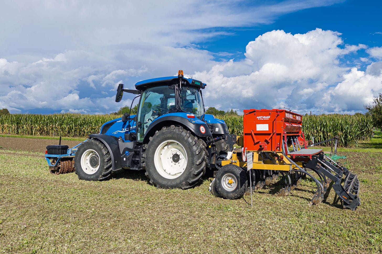 Ein blauer Traktor mit angehängter roter Sämaschine (NT Modell CG300) steht auf einem Versuchsfeld des Oberackers in Zollikofen. Im Hintergrund wächst Mais, während der Vordergrund frisch bearbeitetes Land zeigt. Über dem Feld türmen sich eindrucksvolle weiße Wolken am blauen Himmel.