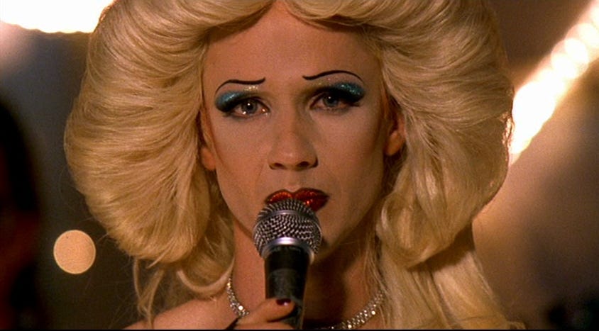 Close-up of Hedwig from the cult film Hedwig and the Angry Inch (2001), wearing a blonde wig, bold blue eyeshadow, and red lipstick while singing into a microphone. The image symbolizes identity as performance, imperfection as leverage, and authenticity through spectacle — themes central to drag culture and personal branding.