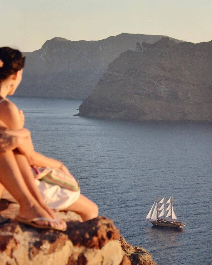 This may contain: a woman sitting on top of a rock next to a sailboat in the ocean