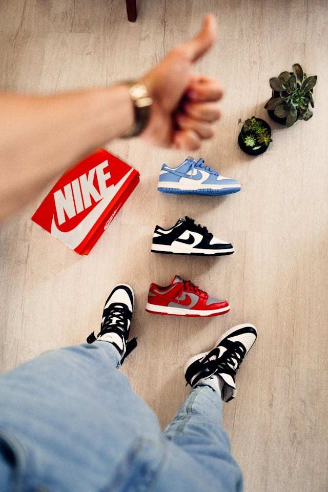 The image shows a flat lay scene featuring Nike sneakers on a light wooden floor. At the center are three Nike Dunk shoes arranged in a display: a light blue and white pair at the top, a black and white pair in the middle, and a red and gray pair at the bottom. In the upper left, a person's hand is giving a thumbs up while wearing what appears to be a gold watch or bracelet. Next to the hand is a red Nike shoebox with the distinctive white swoosh logo. The person is wearing black and white Nike shoes and light blue jeans, with their feet visible at the bottom of the frame. Two small potted succulents can be seen in the upper right corner of the image, adding a touch of greenery to the sneaker collection display. The image shows a flat lay scene featuring Nike sneakers on a light wooden floor. At the center are three Nike Dunk shoes arranged in a display: a light blue and white pair at the top, a black and white pair in the middle, and a red and gray pair at the bottom. In the upper left, a person's hand is giving a thumbs up while wearing what appears to be a gold watch or bracelet. Next to the hand is a red Nike shoebox with the distinctive white swoosh logo. The person is wearing black and white Nike shoes and light blue jeans, with their feet visible at the bottom of the frame. Two small potted succulents can be seen in the upper right corner of the image, adding a touch of greenery to the sneaker collection display.