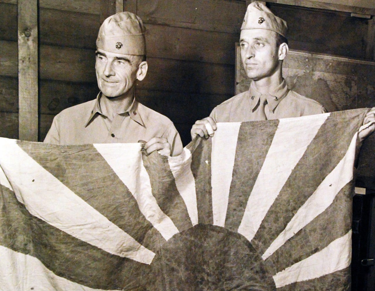 Black-and-white World War II photograph of U.S. Marine LtCol Evans F. Carlson standing beside Major James Roosevelt, both in combat utility uniforms with field gear, holding a captured Japanese flag.