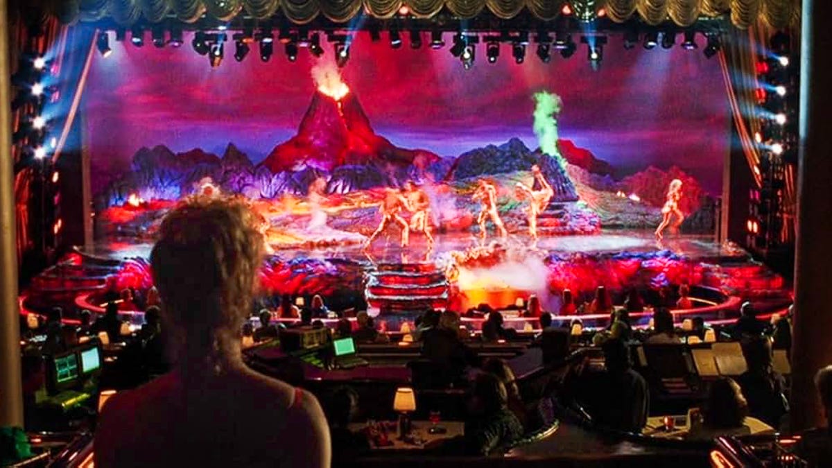 Nomi watches from the back of the theater as scantily clad dancers perform on a volcano-themed stage engulfed in neon lava, smoke, and pyrotechnics, with customers seated at tables in the foreground. Nomi watches from the back of the theater as scantily clad dancers perform on a volcano-themed stage engulfed in neon lava, smoke, and pyrotechnics, with customers seated at tables in the foreground.
