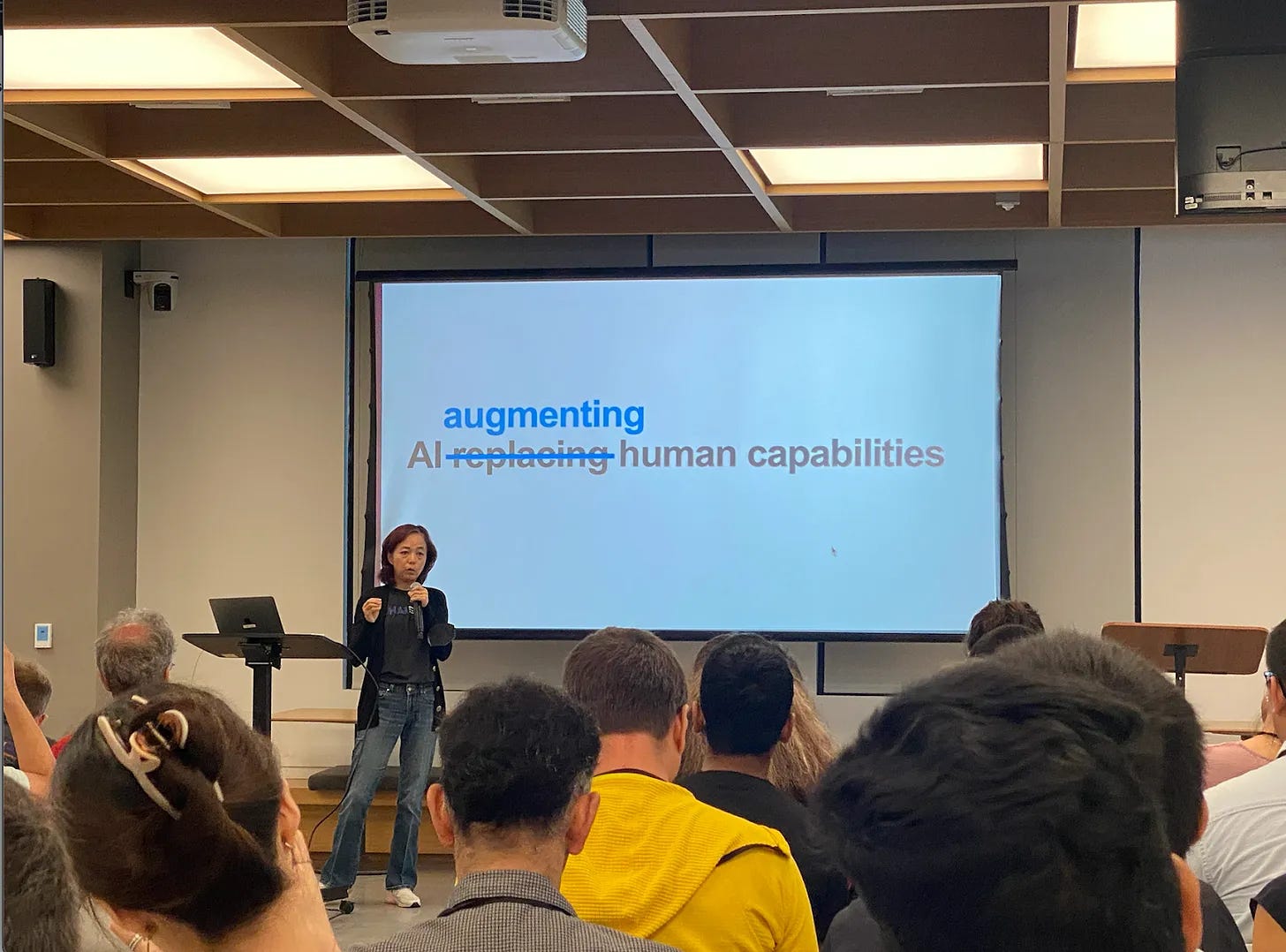 Professor Fei-Fei Li stands in front of a slide that reads "AI augmenting human capabilities". "Replacing" is crossed out, replaced by "augmenting". She is speaking to a packed room. 