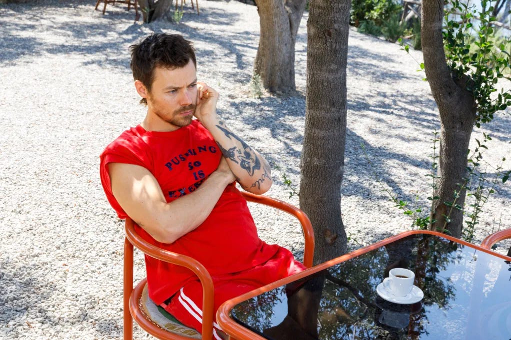 A man with a beard, wearing a red shirt, sits at a table with an espresso cup, in a gravel yard with trees. A man with a beard, wearing a red shirt, sits at a table with an espresso cup, in a gravel yard with trees.