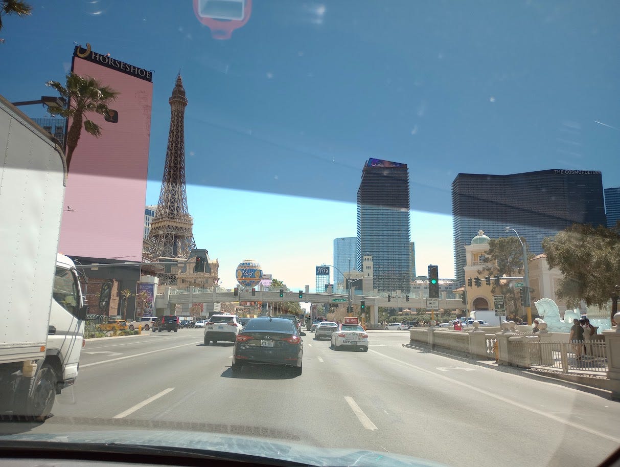 View from car driving up Las Vegas Strip, surrounded by palm trees, tall buildings, and a replica of the Eiffel Tower