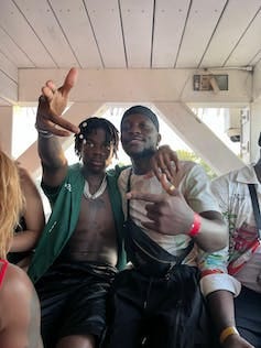Two young African men sit, each with one arm around the other man and the other arm making hand gestures to camera. Two young African men sit, each with one arm around the other man and the other arm making hand gestures to camera.
