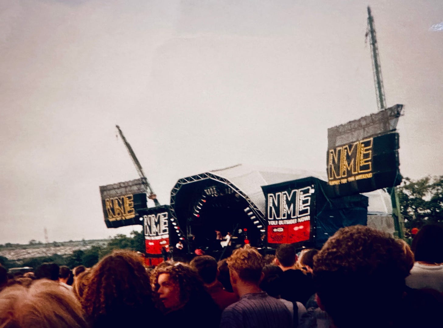 An outdoor stage surrounded by flags that say 'NME' with a large crowd of people in front
