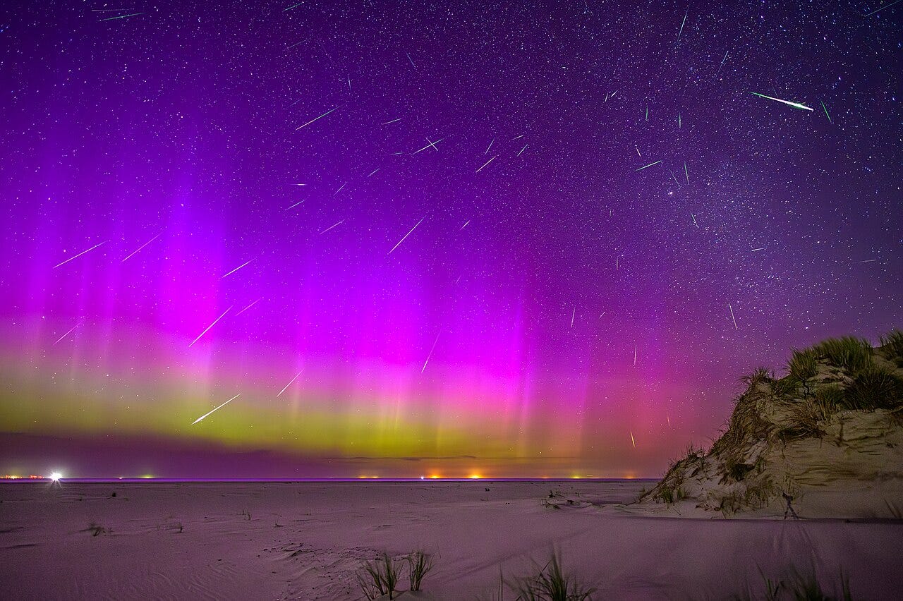 A time-lapse image showing dozens of shooting stars against a yellow, pink, and dark purple sky over a sandy beach