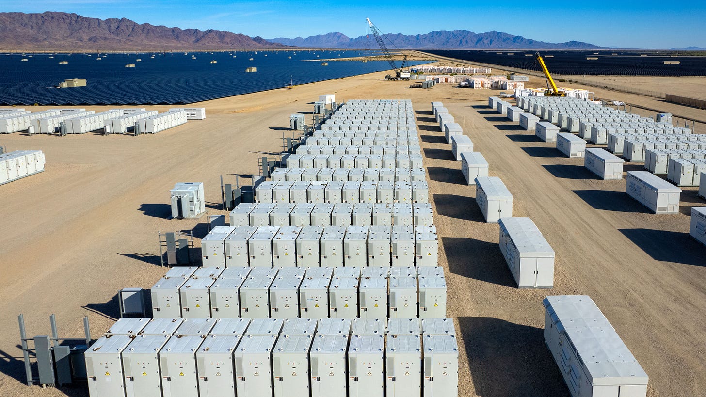 Aerial view of industrial battery units storing electricity in the desert. In the distance are solar panels and mountains.