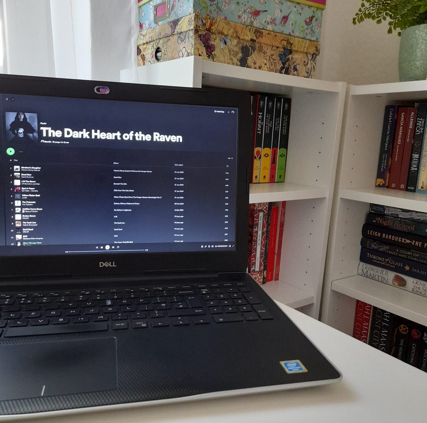 Picture of a laptop with a Spotify playlist called The Dark Heart of the Raven on the screen. In the background are bookshelves.