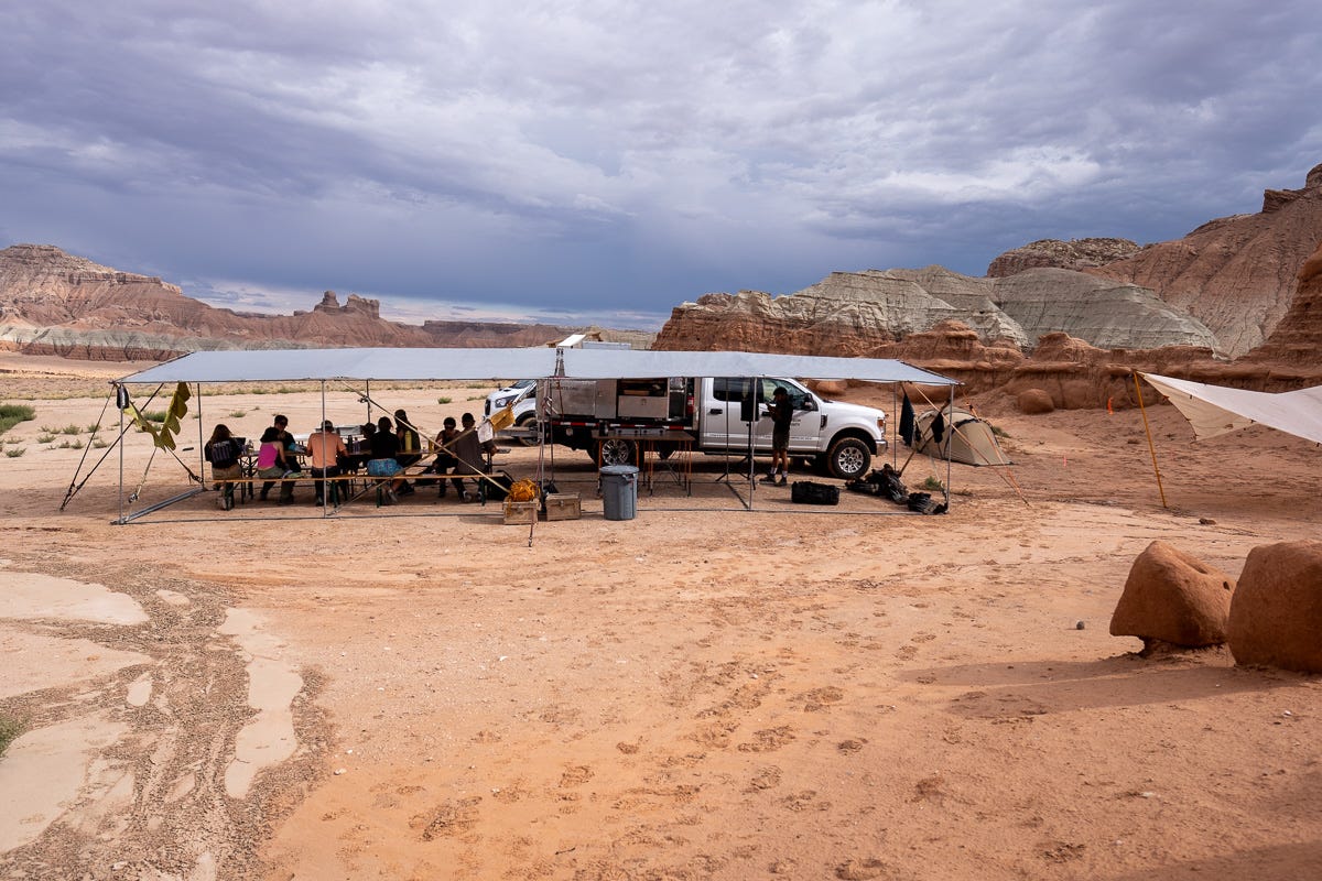 A group of people sit at a long table covered by a tarp structure in front of the Land Arts of the American West truck amidst a sandy red desert landscape with rock formations in the background. A group of people sit at a long table covered by a tarp structure in front of the Land Arts of the American West truck amidst a sandy red desert landscape with rock formations in the background.