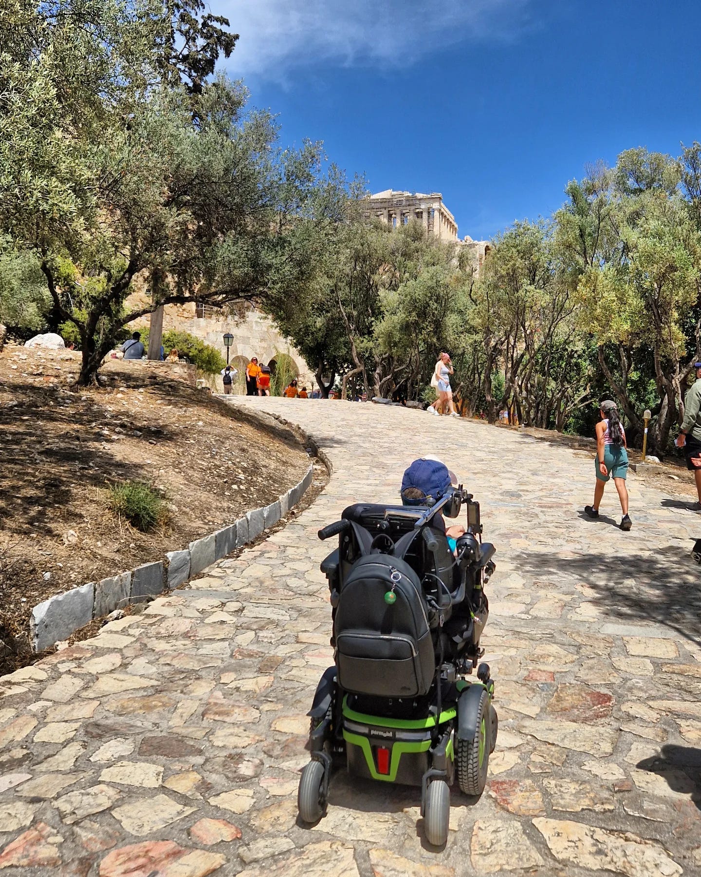 Me rolling up the sloping stone path leading towards the Acropolis. Olive trees line both sides of the route. Several visitors walk ahead and around. The Parthenon is visible at the top of the hill above.