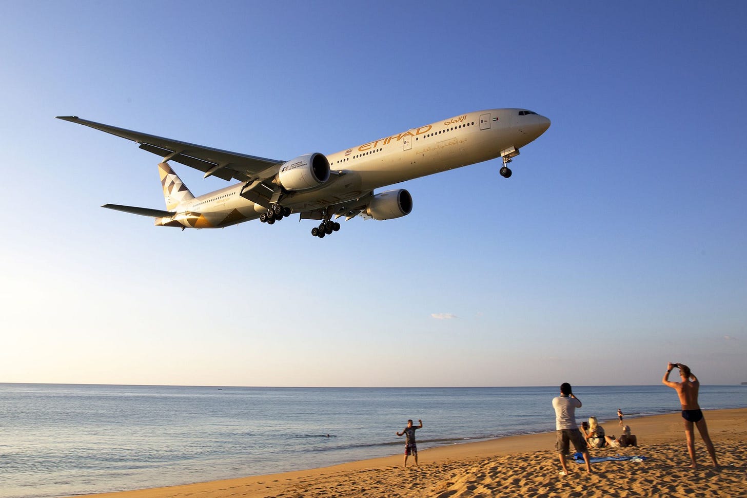 An Etihad Airways airplane flying low over a beach with people watching and taking photos. An Etihad Airways airplane flying low over a beach with people watching and taking photos.
