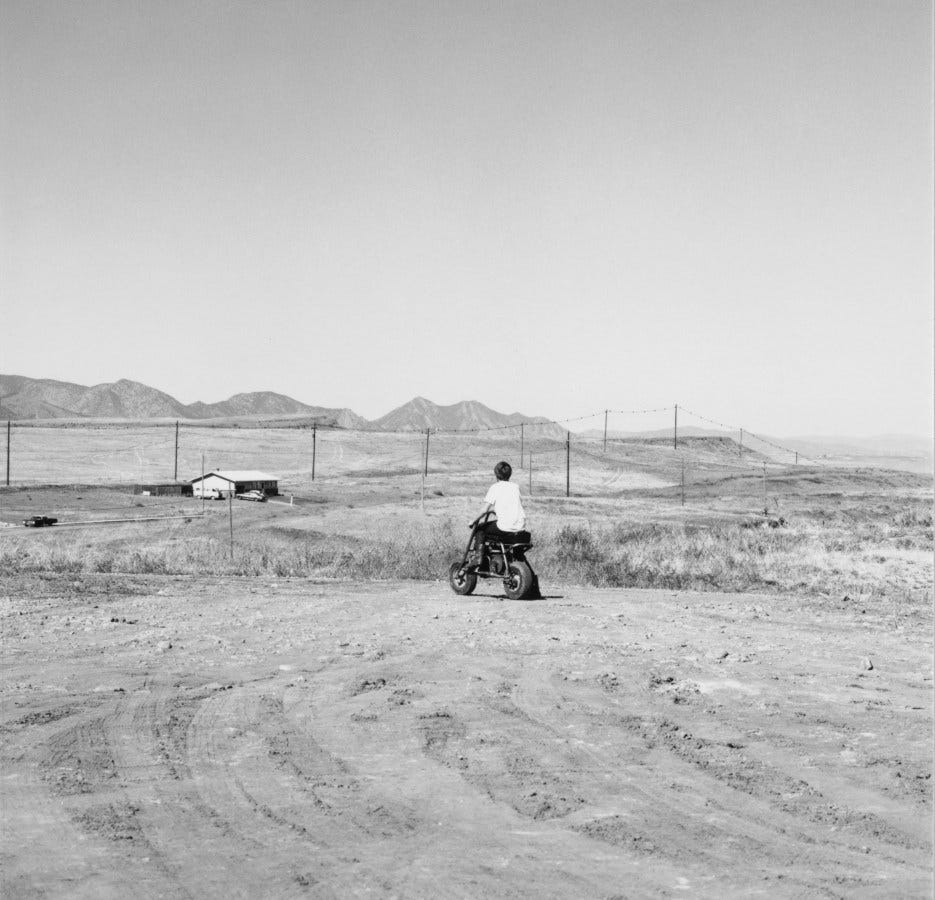 Black and white photograph of a child on a gas powered motorbike admist a dirt field with a house in the distance as well as a mountain range