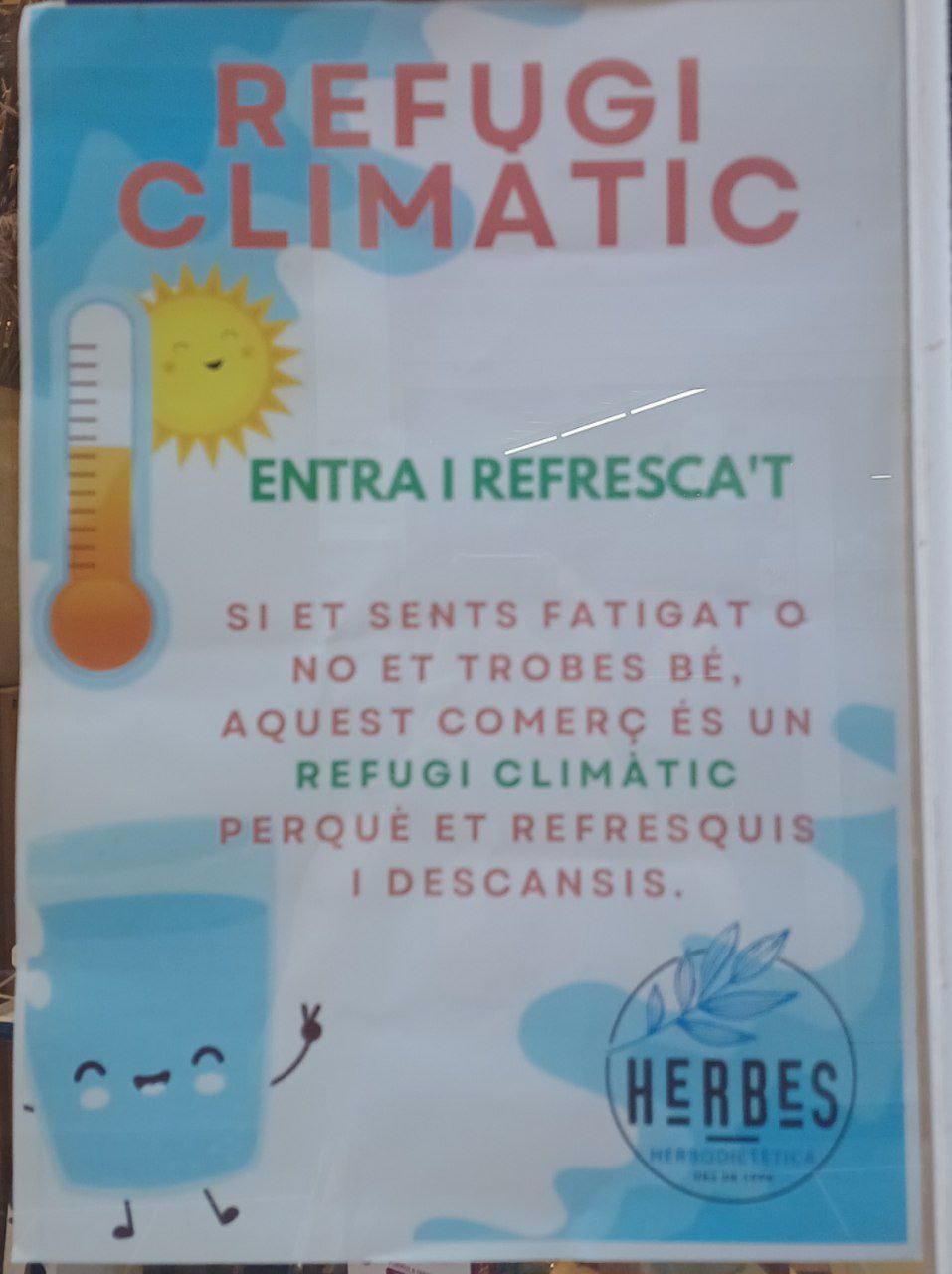 Cartello incollato a una vetrina, illustrato con un sole sorridente, un termometro che misura alte temperature e un bicchiere d'acqua. Il titolo in rosso annuncia in catalano: "Refugi climatic". 