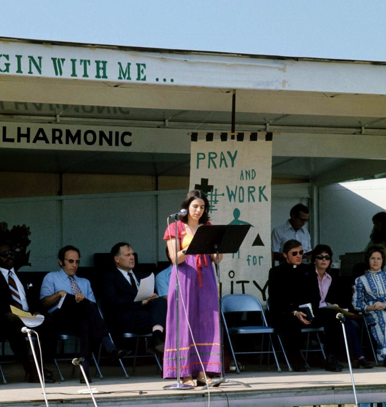 White woman behind podium onstage speaking at 1970s antiwar rally