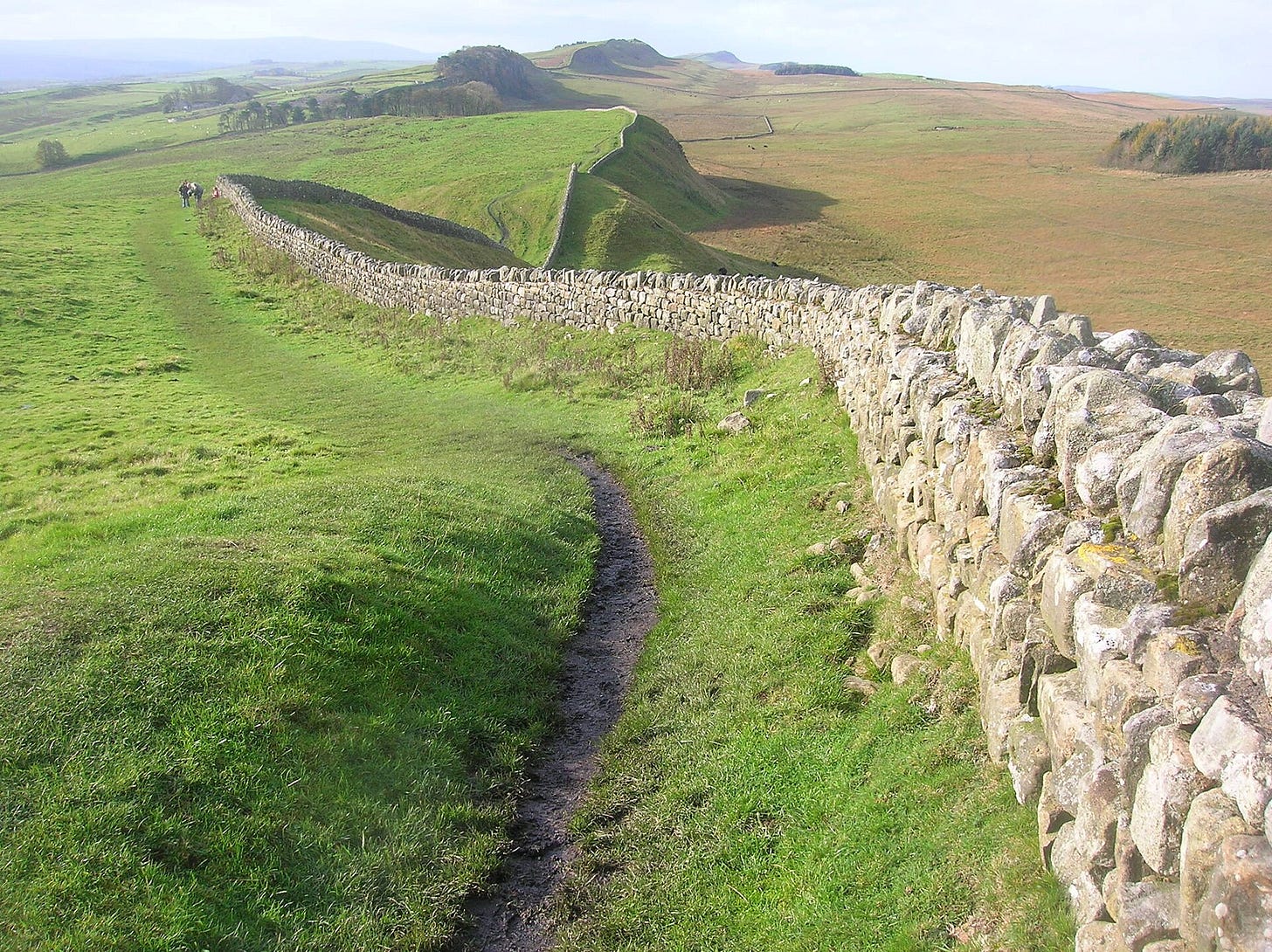 A long, winding section of the ancient stone fortification Hadrian’s Wall at Cuddy's Crag, snaking across the rolling green hills and rugged crags of Northumberland in northern England under a clear sky. A narrow dirt path runs alongside the drystone wall structure on the historic Roman frontier.