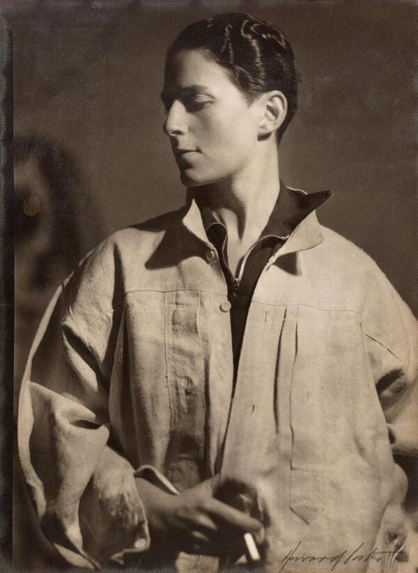 Classic black-and-white portrait of Gluck, a white person, in a loose white shirt/smock, looking down and to the side. Their expression is thoughtful, with shadows adding depth. They have short dark hair, and they are holding a cigarette in their right hand. There is a dark shirt sticking out from under the larger shirt, with both collars sticking up. Classic black-and-white portrait of Gluck, a white person, in a loose white shirt/smock, looking down and to the side. Their expression is thoughtful, with shadows adding depth. They have short dark hair, and they are holding a cigarette in their right hand. There is a dark shirt sticking out from under the larger shirt, with both collars sticking up.