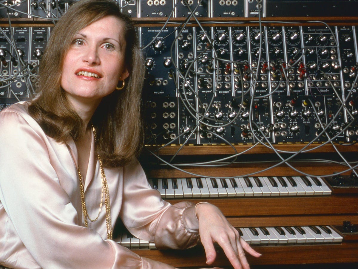 A photograph of a woman in a silk blouse posing in front of a synth machine with two keyboards and numerous electric wires.