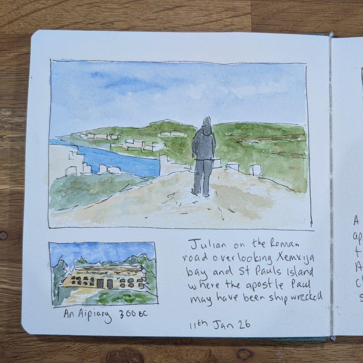 An open sketchbook lies on a wooden table, showing a hand-drawn watercolour scene. The main sketch depicts a lone figure standing on a pale Roman road, seen from behind, looking out over green hills and a blue bay under a soft sky. Below it is a smaller sketch of an ancient stone apiary set into a hillside. Handwritten notes on the page read: “Julian on the Roman road overlooking Xemxija bay and St Paul’s Island where the apostle Paul may have been shipwrecked,” and “An Apiary 300 BC,” dated 11th Jan 26.