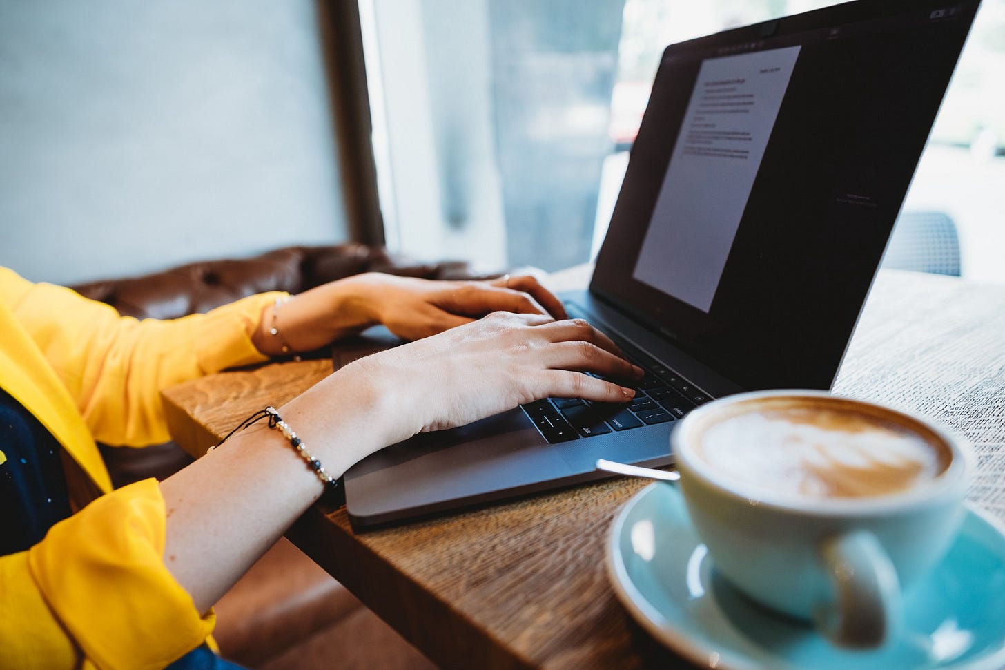 Woman writes on a laptop, a cup of coffee is beside her.