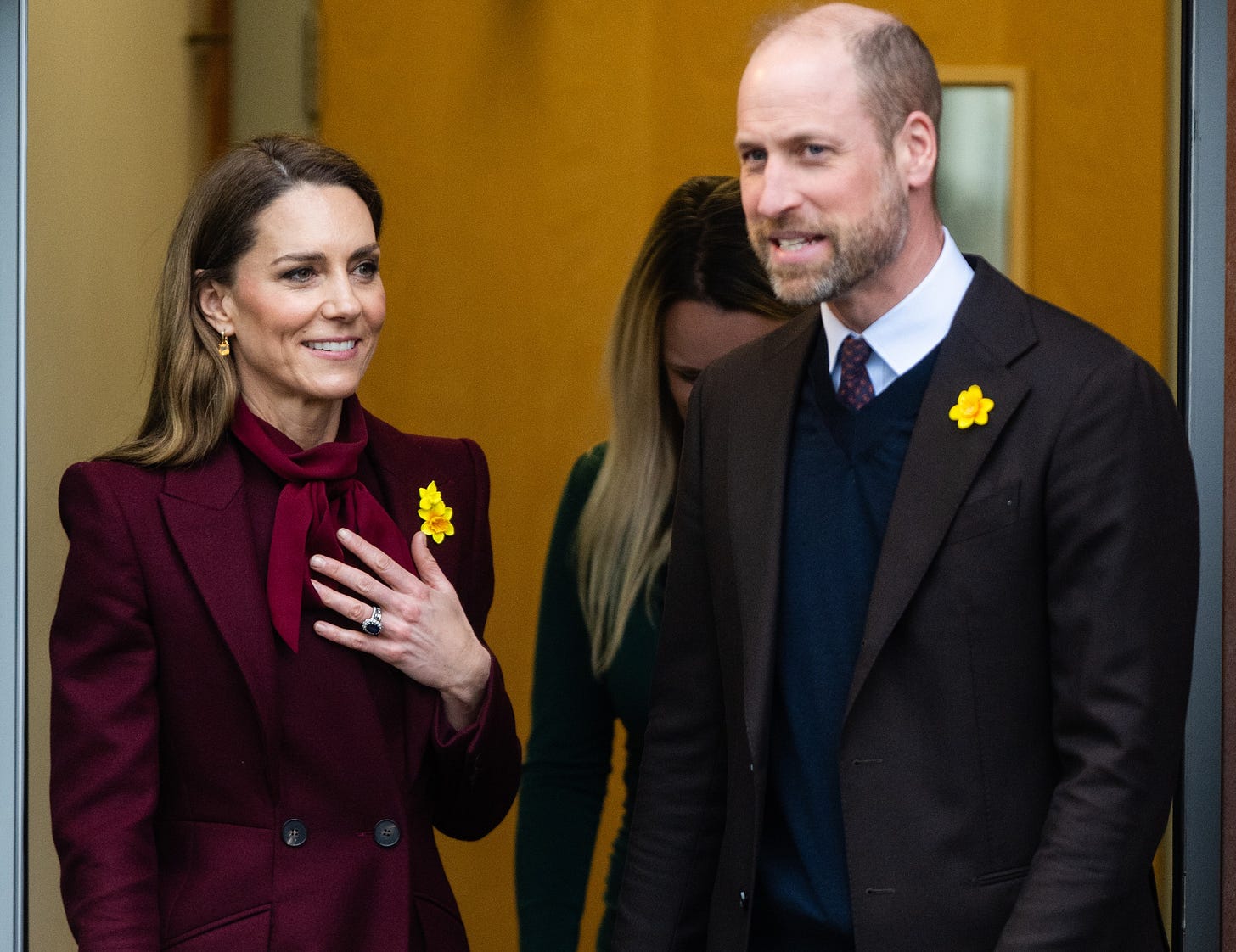 Princess Catherine wears a maroon corduroy suit and smiles alongside Prince William, who is also smiling and wearing a suit. Princess Catherine wears a maroon corduroy suit and smiles alongside Prince William, who is also smiling and wearing a suit.